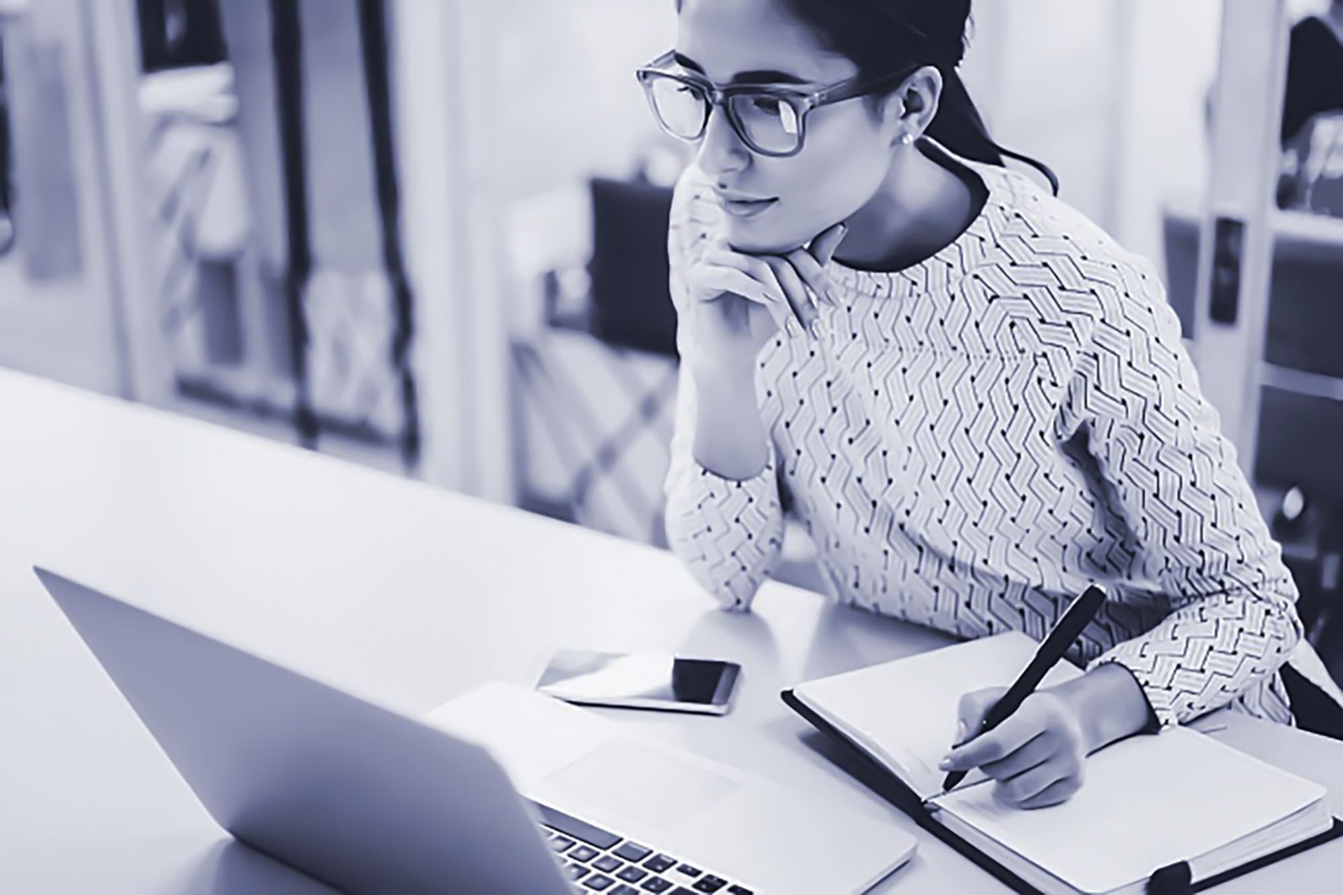 Monochrome image of woman at desk writing in a notebook and looking at a laptop